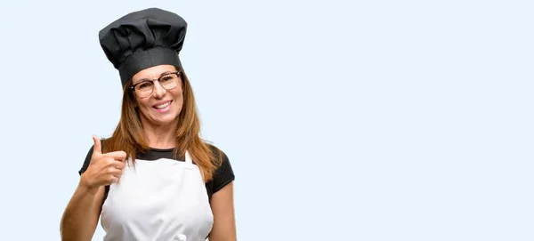 中年厨师妇女穿着厨师围裙微笑广泛显示大拇指向上手势相机 表示喜欢和批准孤立的蓝色背景 — 图库照片
