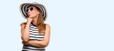 Orta yaş serin kadın yaz şapka ve düşünme ve şüphe ve merak ifade seyir güneş gözlüğü giyiyor mavi arka plan izole