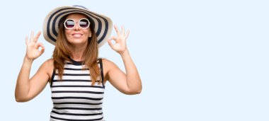 Orta yaş serin yaz şapka ve güneş gözlüğü meditasyon ve gevşeme ifade iki elinle ok işareti jest yapıyor giyen kadın mavi arka plan izole