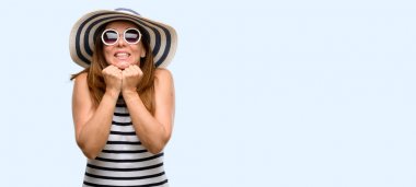 Orta yaş serin yaz şapka ve güneş gözlüğü çok korkmuş ve sinir giyen kadın anksiyete ve panik jest, ifade izole mavi arka plan boğulmuş