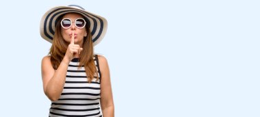 Orta yaş serin yaz şapka ve güneş gözlüğü işaret parmağı ile dudakları, giyen kadın sormak sessiz olmalısın. Sessizlik ve gizli kavramı izole mavi arka plan