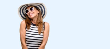 Orta yaş serin kadın yaz şapka ve düşünme ve şüphe ve merak ifade seyir güneş gözlüğü giyiyor mavi arka plan izole