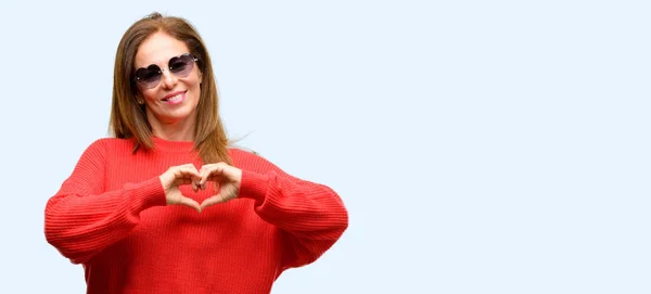 中年妇女戴着心眼镜快乐地显示爱与手在心脏形状表达健康和婚姻符号孤立的蓝色背景 — 图库照片