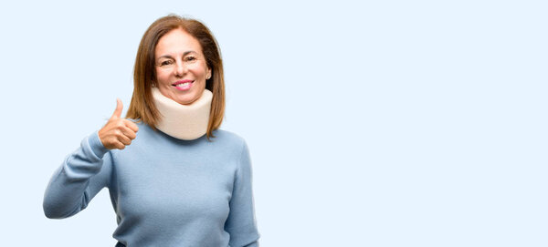 Injured woman wearing neck brace collar smiling broadly showing thumbs up gesture to camera, expression of like and approval isolated blue background