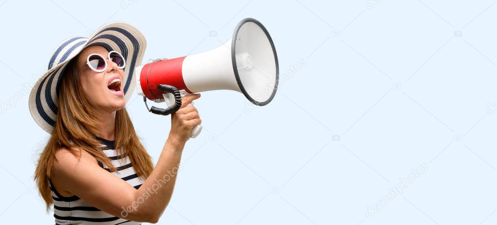 Middle age cool woman wearing summer hat and sunglasses communicates shouting loud holding a megaphone, expressing success and positive concept, idea for marketing or sales isolated blue background