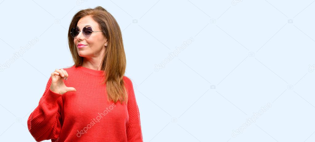 Middle age woman wearing heart sunglasses proud, excited and arrogant, pointing with victory face isolated blue background