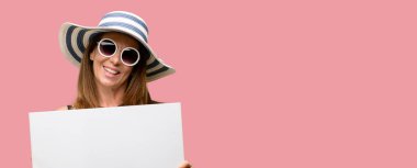 Orta yaş serin yaz şapka ve güneş gözlüğü tutan boş reklam banner, reklam, teklif veya duyuru, büyük kağıt billboard iyi poster giyen kadın