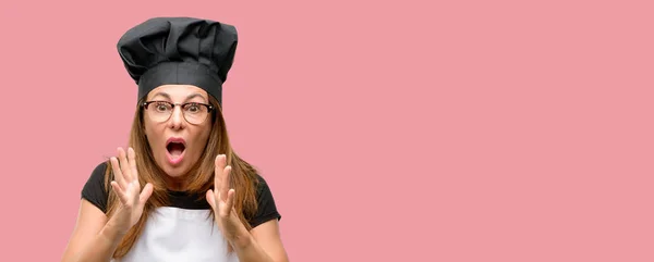 中年厨师妇女穿着厨师围裙压力保持双手头上 吓坏了 在惊慌 — 图库照片