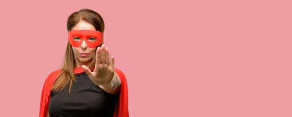 Middle age super hero woman wearing red mask and cape annoyed with bad attitude making stop sign with hand, saying no, expressing security, defense or restriction, maybe pushing