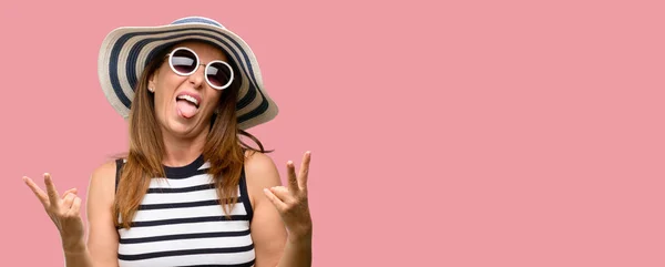 Middle age cool woman wearing summer hat and sunglasses looking at camera showing tong and making victory sign with fingers