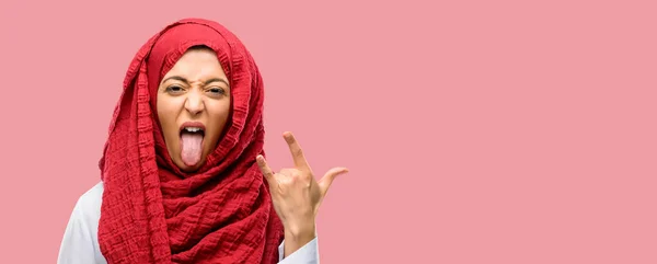 年轻的阿拉伯妇女戴着头巾制作摇滚符号与手 呼喊和庆祝与舌头出来 — 图库照片