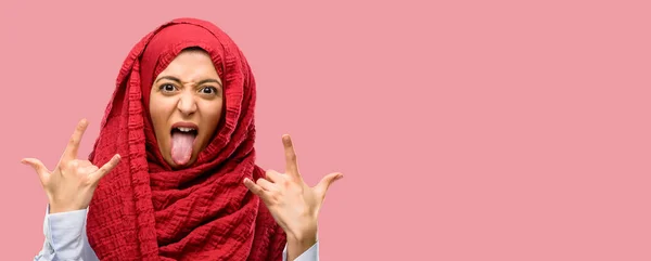 年轻的阿拉伯妇女戴着头巾制作摇滚符号与手 呼喊和庆祝与舌头出来 — 图库照片