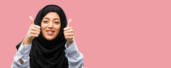 広く示す笑みを浮かべてヒジャーブを着ている若いアラブ女性の親指カメラのような式および承認するジェスチャーを — ストック写真