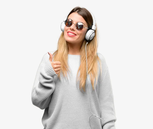 年轻妇女听音乐与耳机微笑宽广地显示竖起大拇指手势对照相机 表示喜欢和批准 — 图库照片