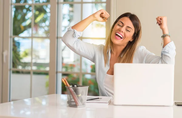 Student woman at table with laptop at home happy and excited celebrating victory expressing big success, power, energy and positive emotions. Celebrates new job joyful
