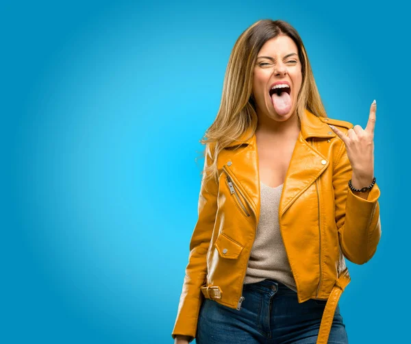 Beautiful young woman making rock symbol with hands, shouting and celebrating with tongue out, blue background