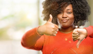 Beautiful african woman confused with thumbs up and down, trying to take a decision expressing doubt and frustration, outdoor clipart