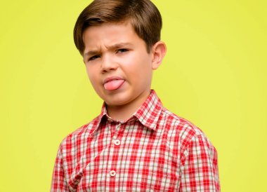 Handsome toddler child with green eyes feeling disgusted with tongue out over yellow background clipart