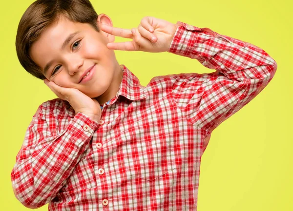 Handsome toddler child with green eyes looking at camera through fingers in victory gesture winking the eye and blowing a kiss over yellow background