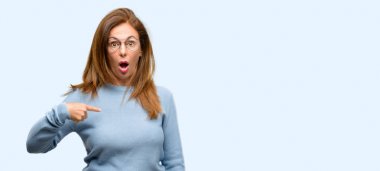 Yün kazak ve serin gözlük mutlu ve tezahürat ifade wow jest, orta yaş kadın parmak izole mavi arka plan ile işaret