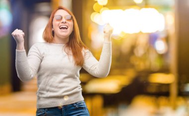 Beautiful young redhead woman happy and excited expressing winning gesture. Successful and celebrating victory, triumphant at night clipart