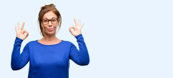 中年の女性はウールのセーターと瞑想とリラクゼーションを表現する両方の手で の手話をやっているメガネを身に着けている分離された青色の背景 — ストック写真