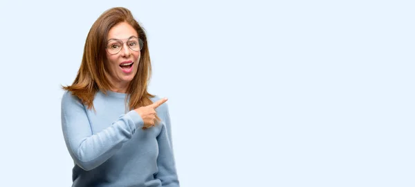 中年の女性はウールのセーターと指でアウェイ側を指しているクールなガラスを身に着けている分離された青色の背景 — ストック写真