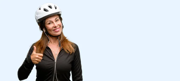 中年サイクリストの女性示す広く笑みを浮かべてイヤホンを使用して親指カメラのような式と分離された承認青い背景にジェスチャーを — ストック写真