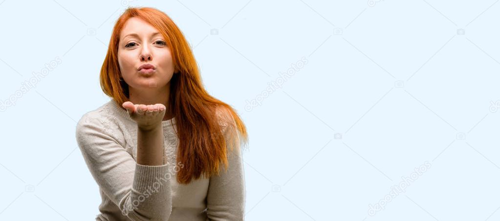 Beautiful young redhead woman expressing love, blows kiss at camera, flirting isolated over blue background