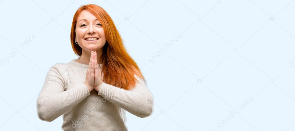 Beautiful young redhead woman with hands together in praying gesture, expressing hope and please concept isolated over blue background