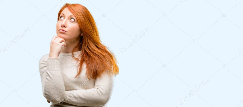 Beautiful young redhead woman thinking and looking up expressing doubt and wonder isolated over blue background