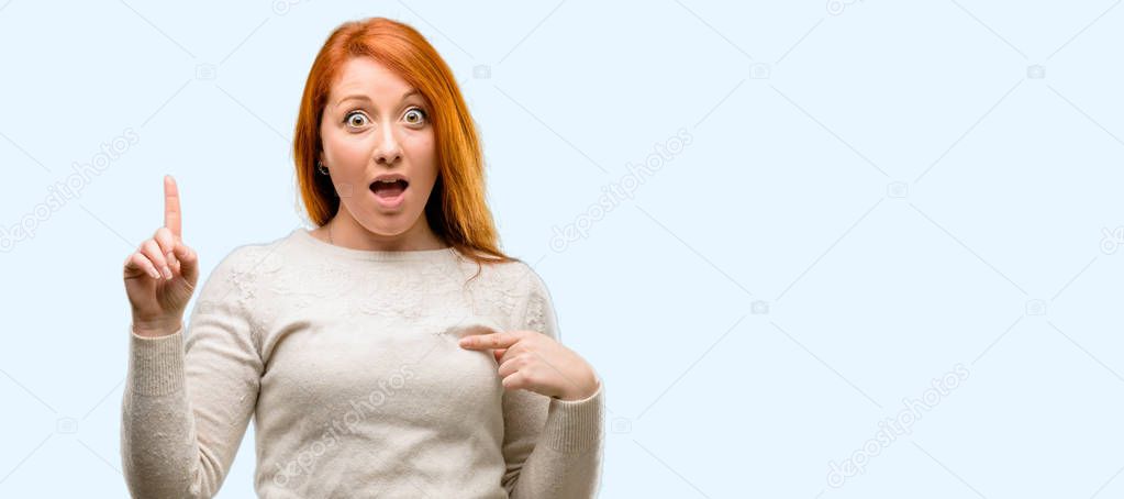 Beautiful young redhead woman happy and surprised cheering expressing wow gesture pointing up isolated over blue background