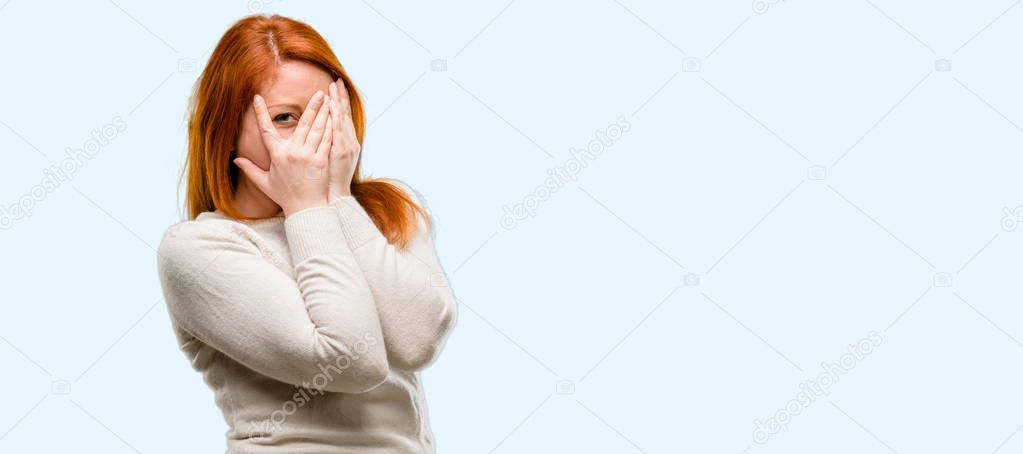 Beautiful young redhead woman smiling having shy look peeking through fingers, covering face with hands looking confusedly broadly isolated over blue background