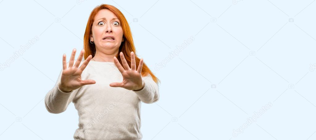 Beautiful young redhead woman disgusted and angry, keeping hands in stop gesture, as a defense, shouting isolated over blue background