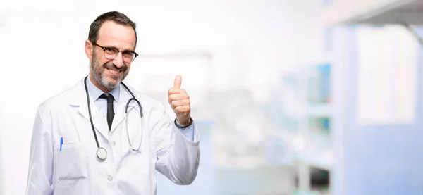 医生资深男士 医学专业的微笑广泛显示竖起大拇指手势照相机 表示喜欢和批准在医院 — 图库照片