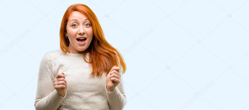 Beautiful young redhead woman happy and surprised cheering expressing wow gesture isolated over blue background
