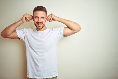 Üzerinde rahat beyaz t-shirt giyen genç yakışıklı adam arka plan gülen çekerek kulakları parmaklar, komik jest ile izole. Seçmelere sorun