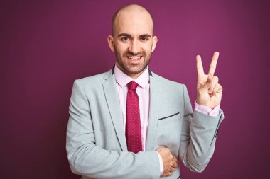 Takım elbiseli ve kravatlı genç bir iş adamı, mor izole edilmiş arka planda kameraya gülümseyen ve zafer işareti yapan mutlu bir yüzle. İki numara..