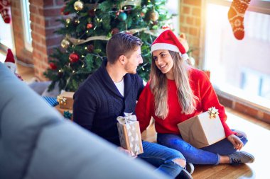 Mutlu ve kendinden emin gülümseyen genç bir çift. Elinde hediyelerle yerde oturup Noel ağacının etrafında sarılmak.