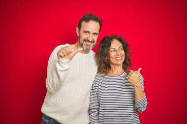 Güzel bir orta yaşlı çift, izole edilmiş kırmızı arka plan üzerine kışlık süveter giyip gülümsüyor ve başparmağıyla yan tarafa bakıyor..