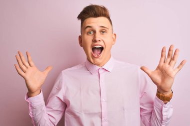 Genç yakışıklı iş adamı zarif gömleğini giymiş pembe arka planda dikilip çılgın ve başarıyı kollarını kaldırıp heyecanla haykırarak kutluyor. Kazanan konsept