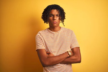 Rastalı tişörtlü Afro bir adam izole edilmiş sarı arka plan üzerinde duruyor şüpheci ve gergin, yüzünde çapraz kollu tasvip etmeyen bir ifade. Negatif kişi.