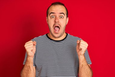 Çizgili donanma tişörtü giyen genç adam izole edilmiş kırmızı arka planda durup kollarını kaldırarak ve açık gözlerle başarıyı kutluyor ve şaşırıyor. Kazanan konsept.