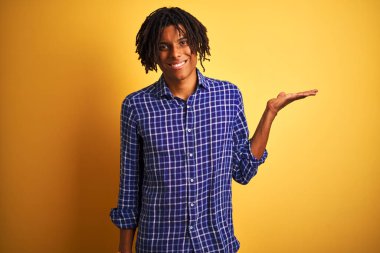 Rastgele gömlek giyen Afro bir adam izole edilmiş sarı arka planda dikiliyor neşeli bir şekilde gülümsüyor ve avuç içi kameraya bakıyor..