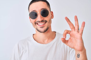 Güneş gözlüğü takan genç yakışıklı bir adam ve izole bir arkaplanda günlük tişört giyiyor. Parmaklarıyla imza atıyor. Mükemmel bir sembol.