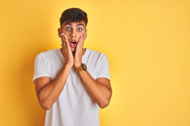 İzole edilmiş sarı arka plan üzerinde duran beyaz t-shirt giyen genç Hintli adam korkmuş ve şok olmuş, şaşırmış ve şaşırmış bir halde