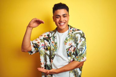 Tatilde olan genç Brezilyalı bir adam yaz çiçekli gömleğini giyip izole edilmiş sarı arka plan jestinin üzerinde büyük ve büyük boyut işareti, ölçü sembolü gösteriyordu. Kameraya bakıp gülümsüyor. Ölçüm konsepti.