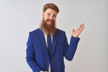 İzole edilmiş beyaz arka planda duran takım elbiseli genç, kızıl saçlı işadamı. Gülümseyen, mutlu bir şekilde elini kaldırıp kameraya bakıyor..