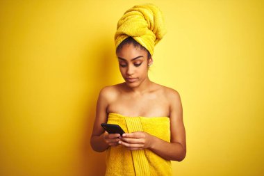 Duş üstüne havlu giyen Afro kadın, izole edilmiş sarı arka plan üzerinde smatrphone kullanıyor. Akıllı bir yüz ifadesiyle ciddi ciddi düşünüyor.