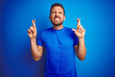Mavi izole edilmiş arka plan jestlerinin üzerine gündelik tişört giyen genç yakışıklı adam umut ve gözler kapalı gülümseyerek el sallıyor. Şans ve batıl inanç..